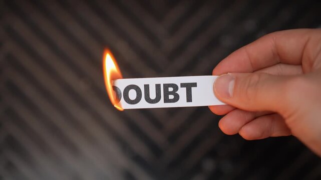 Close-up hand holds a paper strip reading &ldquo;DOUBT&rdquo; burning at the edge, symbolizing overcoming fear, self-belief, and mindset change with copy space on dark background.
