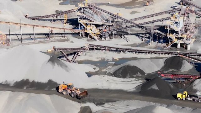 Wheel loader driving across quarry with aggregate processing plant conveyor belt system, active worksite, aerial
