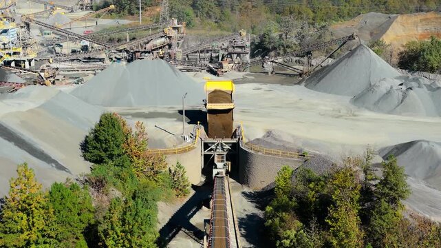 Dump truck unloads fill near gravel mound overlooking hopper and conveyor belt processing plant, aerial pullback