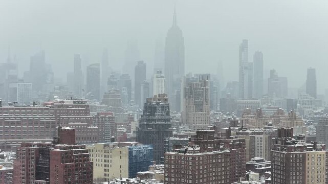 Aerial winter view of Manhattan building blocks covered in snow with skyline and Empire State Building fading into snowy haze.