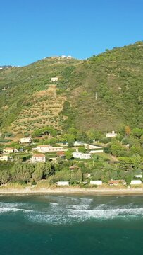 Scenic view of a lush green hillside with houses overlooking the coastline and turquoise sea in Acciaroli, Italy, under a clear blue sky.