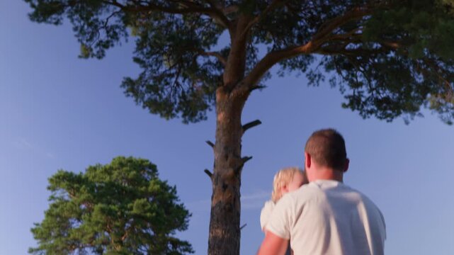 A joyful father with a beard throws his laughing toddler son up in the air. They play in a sunny park with tall pine trees and a clear blue sky, enjoying a happy family moment.