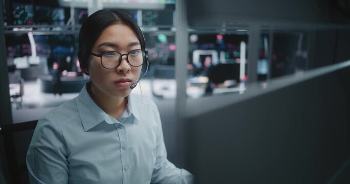 Young Asian Specialist in Crisp Blue Shirt, Focused on Real Time Analytical Dashboards. Woman Wearing Professional Headset For Instant Communication. New Generation Experts in Investment Environment.