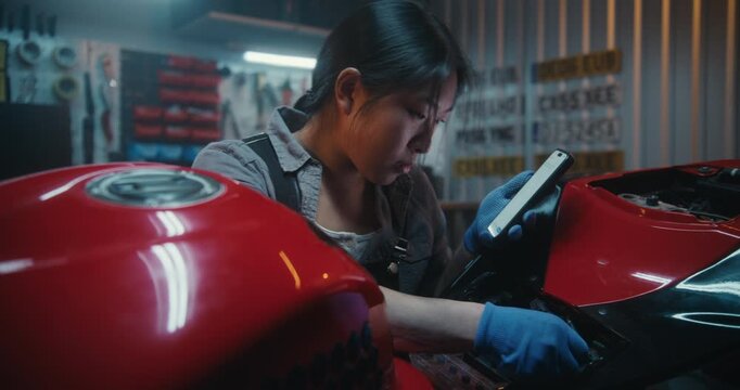 Tech Savvy Female Motorbike Owner Uses Smartphone AI to Diagnose Electrical Issue. Listening to Instructions, Young Woman Attempts Connects Battery and Control Unit Wiring Inside Garage Environment.