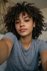 Fototapeta premium Young African American woman with natural curly hair looking thoughtful indoors in casual gray shirt close-up portrait