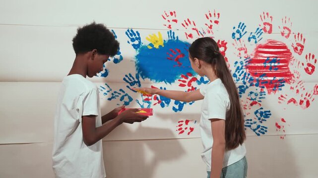 Cute girl and happy african boy put hand in color tray and stamp at stained wall. Diverse high school student painting partition with hand print or doing creative activity in art lesson. Edification