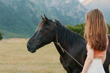 Obraz premium A woman stands beside a dark horse in a wide field with distant mountains. The scene conveys calm companionship and a quiet outdoor bond between human and animal.