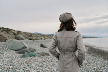 Woman wearing a checkered coat and hat stands on a rocky beach facing the sea under an overcast sky. Concept of autumn fashion, solitude, and coastal landscape. © SHOTPRIME STUDIO