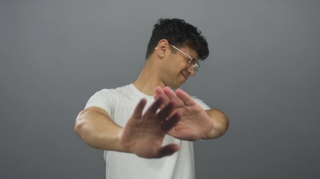 Young hispanic man shielding face with raised hands in blocking gesture against a gray studio wall; denial.