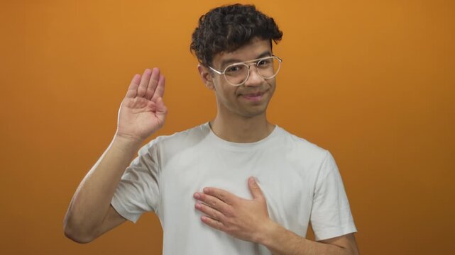 Young hispanic man raises right hand and places left palm on chest in studio with orange background; sincerity oath affirmation.