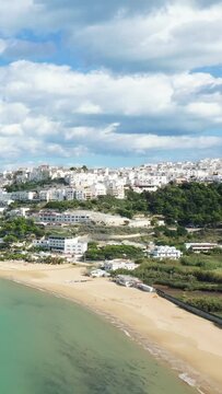 Aerial view of the sandy beach and distinctive white buildings of Peschici, Italy, set against a lush hillside under a partly cloudy sky.