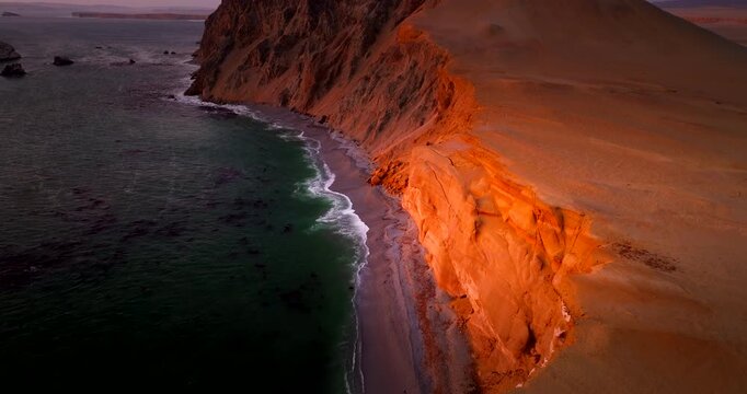 Drone tilts downward over glowing coastal cliffs and beach in Paracas National Reserve, Peru, revealing dark Pacific water and warm desert tones at dusk on South America&rsquo;s coast