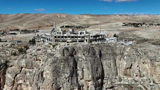 High-angle aerial drone shot of a large stone building perched on the edge of a dramatic rocky cliff in Ma'loula, Syria