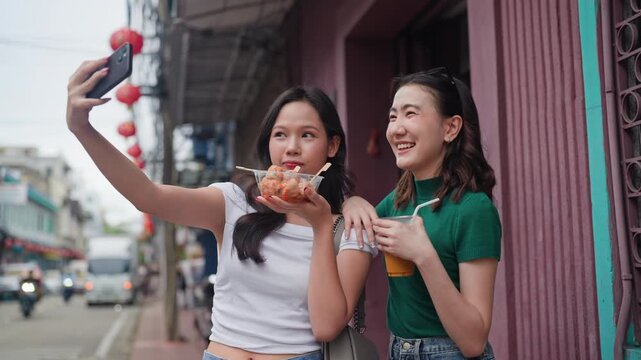 Two joyful Asian women take a selfie on a lively Chinatown street, one holding street food and the other an iced drink, surrounded by red lanterns.