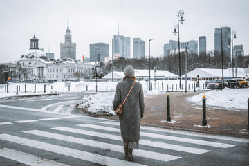 A person walks across a snowy zebra crossing toward a striking urban panorama of Warsaw with the spire of the Palace of Culture and Science.