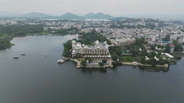 Aerial drone reveals a palatial white-domed hotel on Lake Pichola's shore, with Udaipur cityscape and Aravalli mountains beyond under overcast skies.