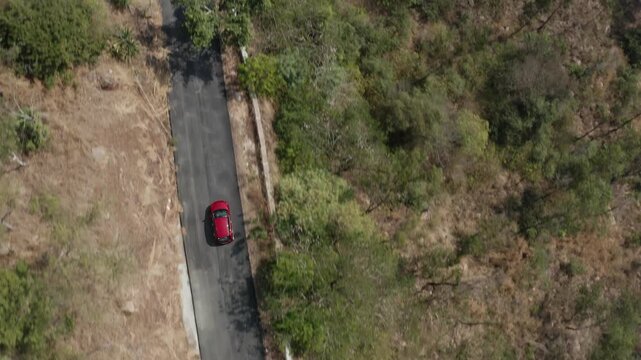 Aerial top-down view of a red car driving along a narrow winding asphalt road cutting through dry hillside terrain on Chamundi Hills near Mysore, Karnataka, with sparse vegetation and strong shadows.
