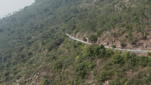 A car navigates the curving asphalt highway winding through dense forest and rocky terrain on the slopes of Chamundi Hill near Mysuru, Karnataka, surrounded by green vegetation and natural boulders.