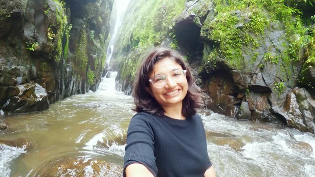 4K Selfie video of Indian teenager girl sitting in front of Satsada waterfall in narrow valley, river stream flowing besides girl. Waterfall background, monsoon landscape. Female tourist recording vlo