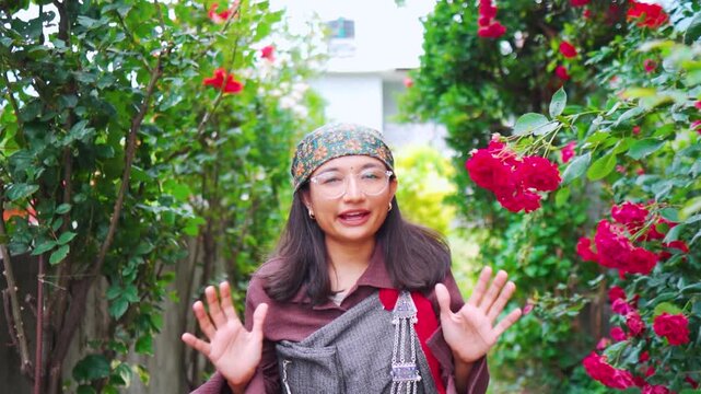 Portrait of Indian woman dressed in traditional Kulluvi dress Pattu and scarf known as Dhatu. Confident woman recording vlog and saying hi, hello in camera. Tribal female standing in rose garden at ho