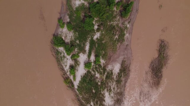 Forward moving downward looking view of island in muddy polluted water of the Makong River, with patches of green foliage, during the rainy monsoon season.