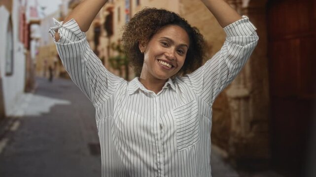 Woman raising arms and smiling on a narrow cobbled street in old town while looking upward with open hands; joy wander curiosity.