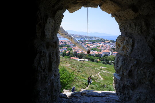 A scenic view of the coastal town of Foca with red-roofed houses and the blue sea, framed by the rough stone window of a historic windmill in Turkey