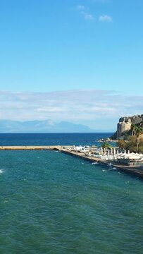 Scenic view of a coastal promenade and harbor with clear blue water and distant mountains in Koroni, Greece. Sunny weather with a vibrant, inviting atmosphere.