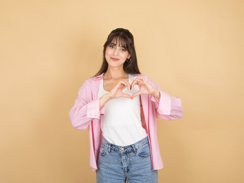 A charming young woman making a heart shape with her hands over her chest while smiling. She is dressed in a pink open shirt and denim jeans against a neutral beige studio background.