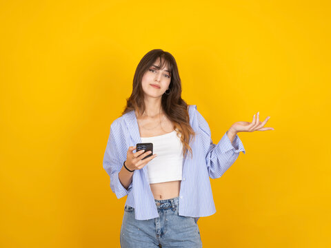 A confused young woman holding a smartphone and gesturing with an open hand against a vibrant yellow background. She wears a striped blue shirt over a white crop top and denim jeans.