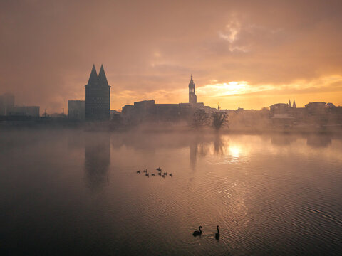 Aerial view of a misty sunrise casting golden hues over the water reflecting the silhouette of Munsterkerk and St. Christopher Cathedral, Roermond, Limburg, Netherlands.