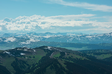 Fototapeta premium Aerial sunlit top view above forest hills and big snowy mountain range under lush clouds in changeable weather. Scenic hilly alpine landscape with sunlight and shadows. Partly cloudy in high mountains