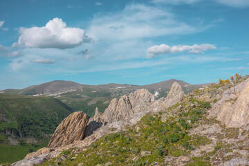 Fototapeta premium Green grasses and rock outcrops on cliff above alpine valley against hills and mountains far away under clouds in blue sky. Sharp rocky outliers on precipice edge in bright sun in cloudy sunny day.