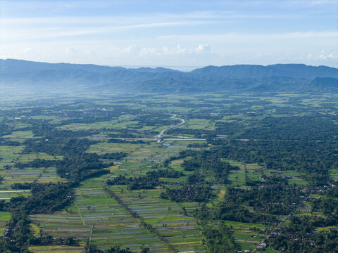 Aerial view of the meandering Woro River cutting through a vibrant green landscape beneath a hazy sky, Klaten Regency, Central Java, Indonesia.