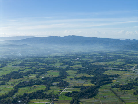Aerial view of a lush, green landscape woven with rivers, under a hazy sky meeting distant mountains, Woro River, Klaten Regency, Central Java, Indonesia.