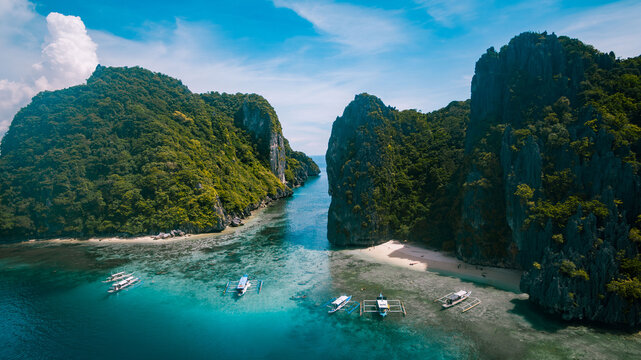 Aerial view of turquoise waters meet towering limestone cliffs, fringed by emerald foliage and pearl-white beaches, El Nido, Mimaropa, Philippines.