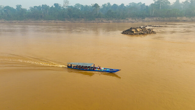 Aerial view of a boat cuts through the muddy river, a vibrant contrast against the dense green rainforest along the banks, Puerto Maldonado, Madre de Dios, Peru.