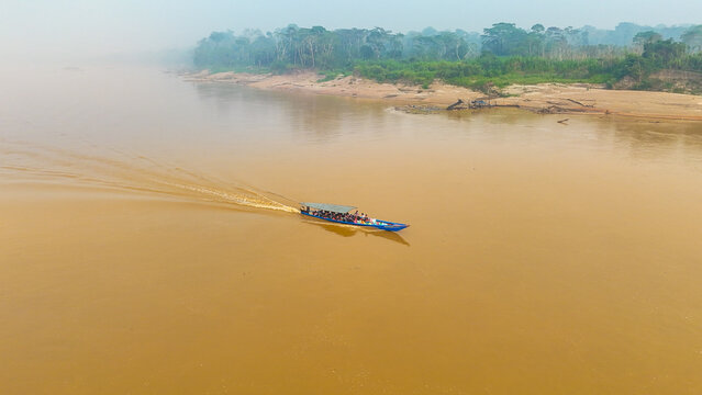 Aerial view of a boat slicing through the muddy waters of the Madre de Dios River, framed by the hazy, verdant rainforest, Puerto Maldonado, Madre de Dios, Peru.