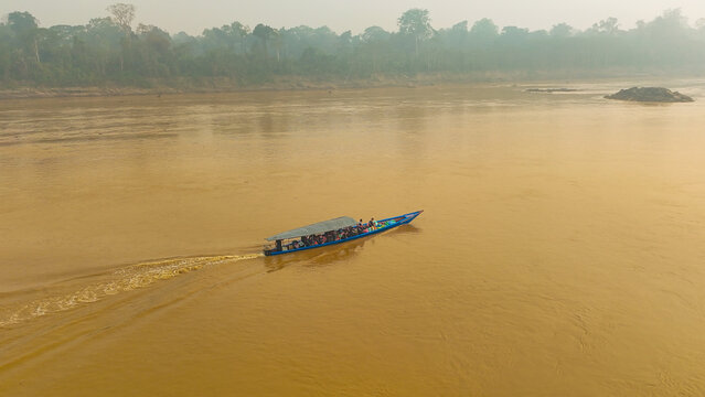 Aerial view of a small boat cutting through the murky river waters, framed by the distant green of the Amazon rainforest, Puerto Maldonado, Madre de Dios, Peru.