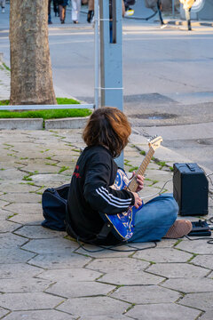 Street musician sitting on a hexagonal paved sidewalk, playing a blue electric guitar with a portable amplifier.