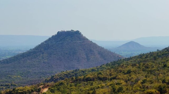 Distant scenic view of Trikuta Hill with Maa Sharda Temple at the summit surrounded by forested hills and layered countryside landscape under hazy blue sky.
