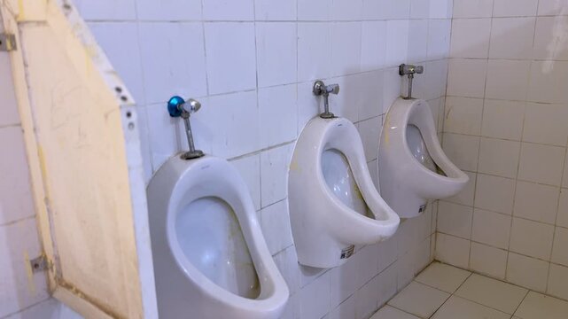 Side view of three wall mounted ceramic urinals with visible stains and divider panel inside an aging tiled public restroom showing sanitation fixtures and indoor plumbing setup.