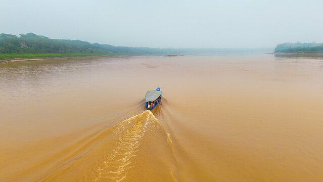 Aerial view of a boat slicing through the muddy waters of a river, with lush green forest lining the banks under a hazy sky, Puerto Maldonado, Madre de Dios, Peru.