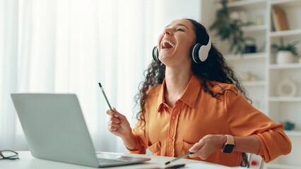 A woman sits at a desk in her home, wearing headphones and smiling while listening to music. She...
