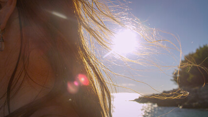 CLOSE UP, LENS FLARE: Gentle wind lifts her long brown hair, which shimmers in the warm sunlight on the sea. Water and shoreline sit softly in the background, creating a peaceful summer holiday mood.