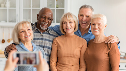 A group of five older adults stands close together in a kitchen. They smile and pose for the camera while one person takes their picture. The kitchen is bright and has many cabinets. © Prostock-studio
