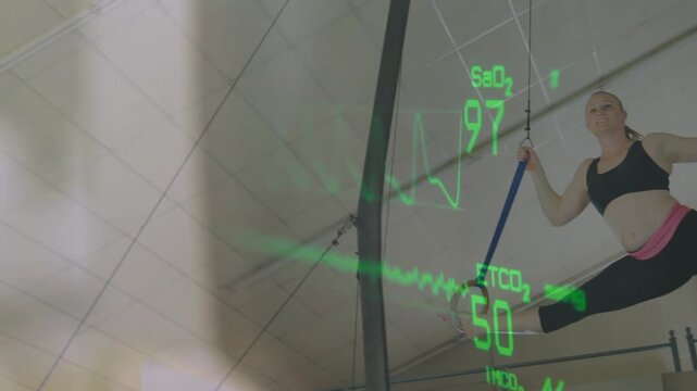 Female gymnast gripping straps, pulling into rings in gym while fitness overlay tracking SaO2 or ETC