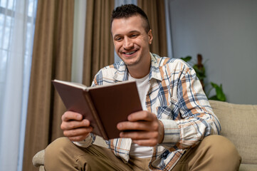 Joyful man holding book seated on sofa in living room interior