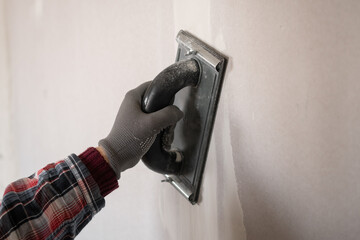 Close-up of worker hand in orange glove sanding plaster on drywall using a plaster hand tool for...