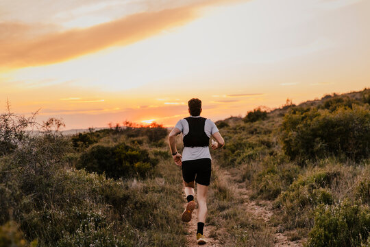Trail runner running along a narrow mountain path toward a colorful sunset horizon. Concept of endurance sports, adventure running and outdoor lifestyle
